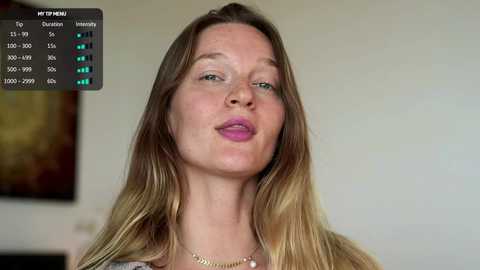 Video of a Caucasian woman with long, blonde hair, wearing pink lipstick, smiling, with a background showing a blurry, modern painting and a digital screen displaying her profile details.