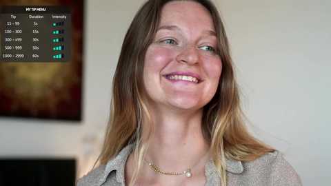 Video of a smiling Caucasian woman with long blonde hair, wearing a gray cardigan, standing indoors. Background shows abstract art and a digital display with stats.