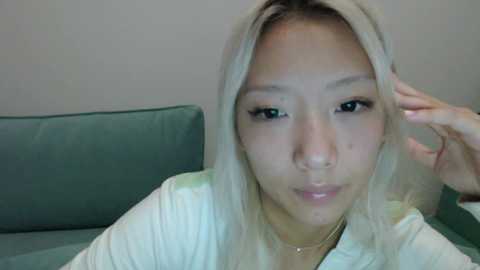 Video of an East Asian woman with long, straight, platinum blonde hair, light skin, and almond-shaped eyes, wearing a white shirt, resting her hand on her head, against a green couch.