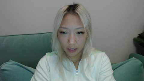 A video of an Asian woman with shoulder-length platinum blonde hair and light skin, wearing a white jacket, sitting on a green couch against a plain white wall.