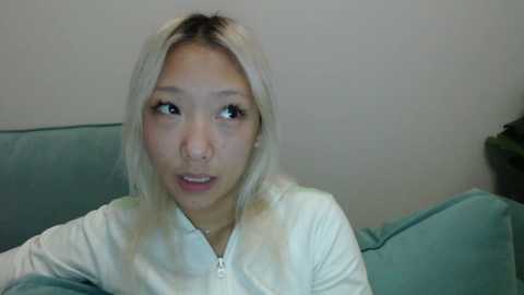 Video of a young Asian woman with long, straight, platinum blonde hair, wearing a white zip-up hoodie, looking surprised with a hand on her cheek. She sits on a teal couch against a plain white wall.