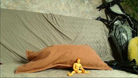 Video of a plush Jack Skellington doll lying on a beige quilt, with a yellow figurine on the floor, set against a textured, pale green wall.