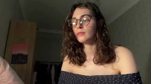 Video of a young woman with curly brown hair, wearing glasses, a dark off-shoulder top, and a neutral expression. Background features a dimly lit bedroom with a wooden door and a partially visible closet.