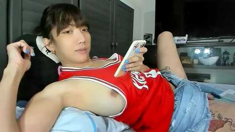 Video of a young Asian woman with short black hair, wearing a red tank top with white text and denim shorts, lying on a bed, playing with a phone, in a dimly lit bedroom.