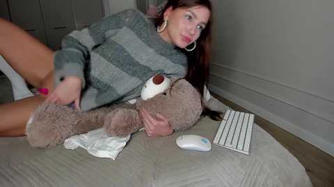 Video of a woman with long brown hair, wearing a striped sweater, lying on a bed with a stuffed animal, white mouse, and gray blanket.