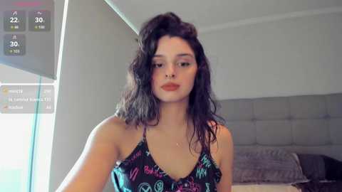Video of a young woman with long, wavy dark hair, wearing a colorful, low-cut top, standing in a modern, softly lit bedroom.
