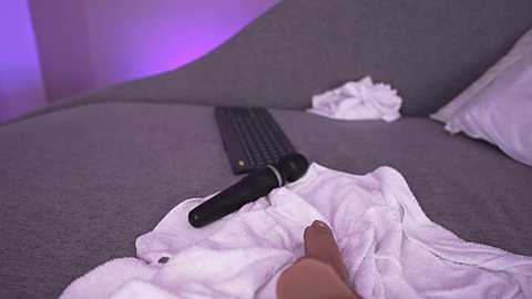 A video of a person reclining on a gray couch, with a white blanket and a black vibrator, surrounded by soft purple lighting, creating a cozy and intimate atmosphere.