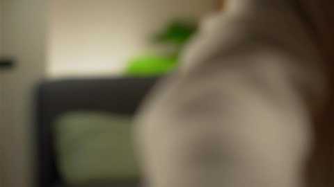 A blurred video of a naked person with pale skin, standing in a dimly lit room with a green plant in the background.