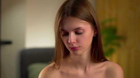 Video of a young Caucasian woman with straight brown hair, wearing minimal makeup, standing topless in an indoor setting with a blurred background.