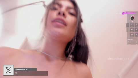 A video of a topless woman with long dark hair, fair skin, and a serene expression, taken from a low angle, showing her chest and shoulders. The background is a blurred white wall.