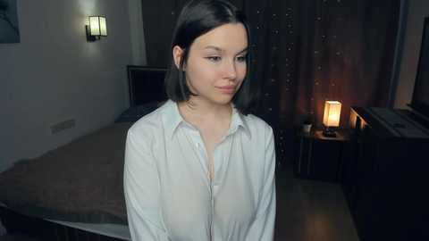 Video of a young woman with straight black hair, wearing a white button-up shirt, standing in a dimly-lit bedroom with a bed, nightstand lamp, and dark curtains.