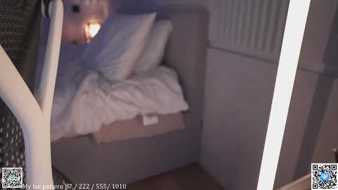 A dimly lit, blurry video of a cozy bedroom with a white bed, white pillows, and a nightstand with a lit lamp. A person's arm and part of their face are visible on the left.