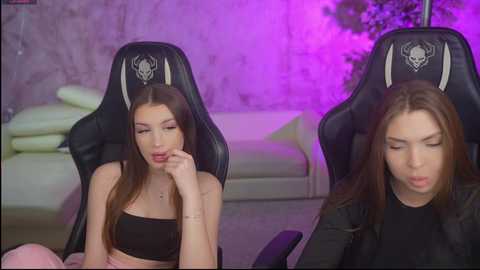 Video of two young women with long hair, one in a black crop top, the other in a black jacket, seated in black gaming chairs, under purple lighting in a room with a white sofa.