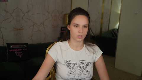 Video of a young woman with dark hair, wearing a white t-shirt with \"Always Forget\" printed on it, seated indoors with a dark green couch and a cluttered background.