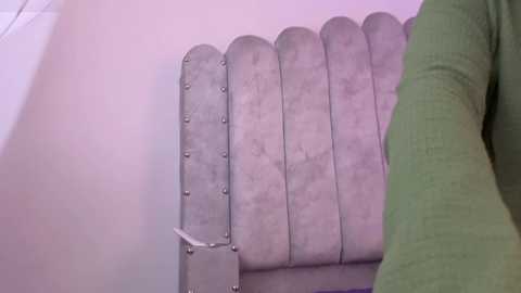A video of a person's arm in a green long-sleeve shirt, resting on a light gray, tufted bench with metallic buttons and a textured, slightly worn fabric. The background is a light pink wall, creating a soft, pastel color palette.