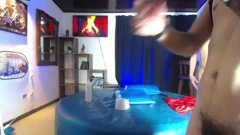 Video of a dimly lit, themed room with blue inflatable beds and red sheets. Wall-mounted fireplace and shelves with motorcycle-themed items. Man in motion, wearing a black hat, visible from the waist up.