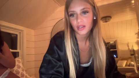 Media: Video of a blonde woman with straight hair, wearing a black jacket, sitting indoors in a cozy room with wooden walls and a TV.