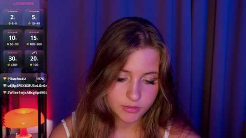 Video of a young woman with light skin and long brown hair, wearing a white top, against a purple curtain background. She appears to be on a live stream with various social media icons displayed.