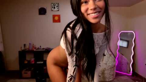 A video of an Asian woman with long black hair, smiling, wearing a cropped white shirt, leaning over, in a dimly lit room with neon lights, shelves, and artwork.