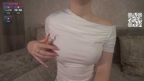 A video of a woman with fair skin, wearing a tight, white off-shoulder top, with large breasts, partially visible nipple, and a ring on her left hand, seated on a gray couch.