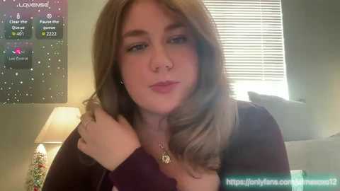 Video of a young Caucasian woman with shoulder-length blonde hair, wearing a dark top, standing indoors near a window with blinds. The background shows a TV screen displaying \"Love Island\" content.