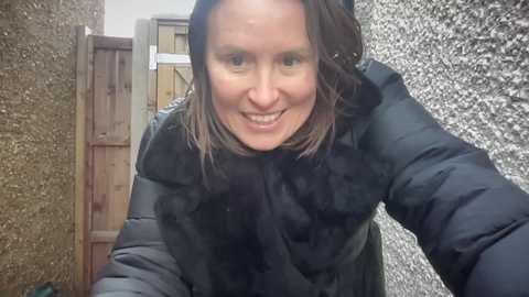 Video of a smiling woman with shoulder-length brown hair, wearing a black puffy coat, standing in a rustic outdoor setting with a wooden fence and textured stucco wall.