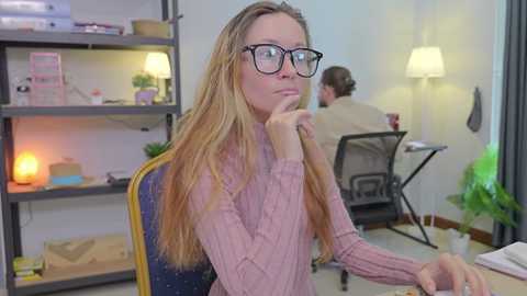 Video of a blonde woman with long hair, wearing glasses, a pink sweater, and holding her chin, seated at a desk in a modern office with shelves, green plants, and a lit lamp.