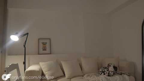 A video of a minimalist, beige-walled living room featuring a plush, cream-colored sofa with white cushions, a leopard-print cushion, and a floor lamp with a white shade. A framed abstract painting hangs on the wall.