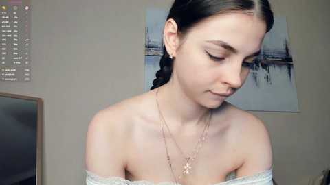 Video of a young woman with fair skin, dark hair in a braid, wearing off-shoulder lace top and multiple layered necklaces. She looks down with a serene expression in a beige room with a framed painting of a river scene.