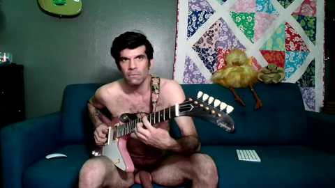 Video of a shirtless man with short dark hair playing an electric guitar on a teal couch. Background features a colorful quilt and a stuffed toy duck.