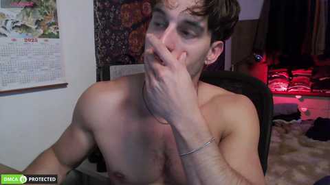 A video of a shirtless, pale-skinned man with messy brown hair, covering his mouth with his hand, in a dimly lit room with a calendar, art, and red lighting.