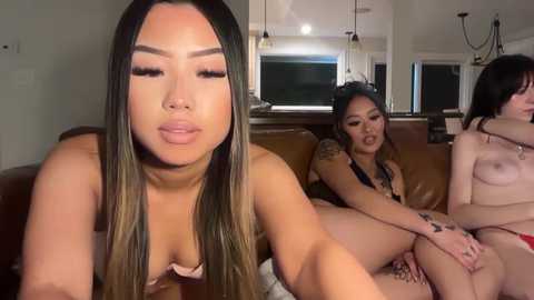 Video of two Asian women, one topless, on a brown couch, with a third partially visible, in a modern kitchen with white cabinets and windows.