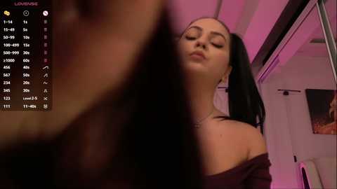 Video of a young woman with long black hair in pigtails, wearing a dark off-shoulder top, standing in a dimly lit room with pink lighting.