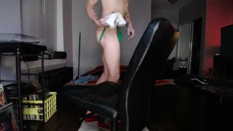 Video of a slender, fair-skinned woman in a green thong, holding a white cloth, standing in a cluttered, dimly lit room with a black chair, a desk, and a bed.