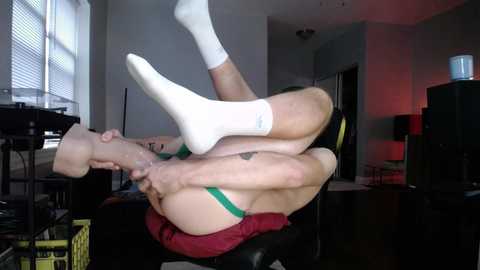 Video of a man in white socks and green underwear, lying on his back, legs bent, and feet touching his face in a dark room with a window, a black speaker, and a desk.