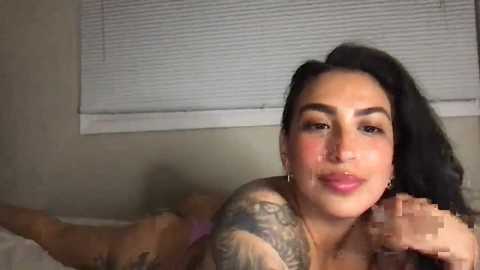 Video of a Latina woman with long black hair, tattoos, and a full face of makeup lying on a bed, partially obscured by a man's hand.