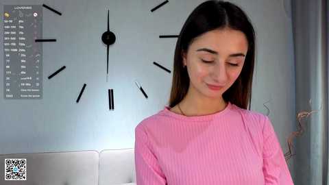 Video of a young woman with straight dark hair, wearing a pink ribbed sweater, standing in a minimalist room with a large wall clock and white couch.