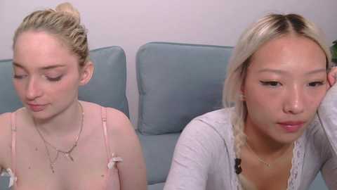 Video of two young women with fair skin and blonde hair, seated side-by-side on a light blue couch. One wears a pink dress, the other a white cardigan. Both appear tired, with the blonde woman resting her head on her hand.