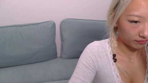 Video of an East Asian woman with long, blonde braided hair, wearing a white, long-sleeved top, sitting on a light blue couch against a plain white wall.