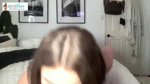 Video of a woman's back, showing light brown hair with a visible part, lying on a bed with white and black sheets. White door with towels hanging behind.