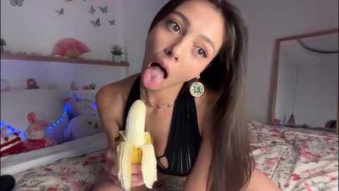 Video of a young Asian woman with long brown hair, wearing a black halter top, licking a peeled banana in a colorful bedroom with butterfly wallpaper.