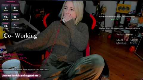 Video of a blonde woman in a gray sweater, sitting on a red gaming chair, wearing a headset, surrounded by gaming peripherals in a dimly-lit room.
