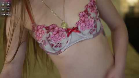 Video of a slender, light-skinned woman with long brown hair, wearing a pink and white floral bra with red bows, and a gold pendant necklace. Background is blurred, focusing on her torso and upper chest.