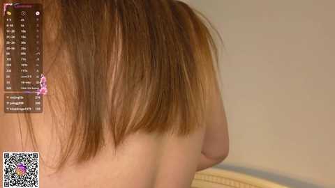 Video of a woman's upper back and shoulder, showing a straight, shoulder-length brown bob haircut. In the background, a QR code and a digital clock display are visible. The setting appears to be an indoor environment with neutral-colored walls.