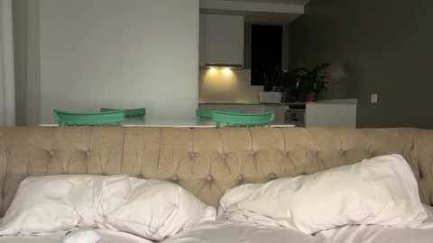 Video of a modern, minimalist bedroom with a beige, tufted headboard, two green chairs, and a small kitchen visible in the background.
