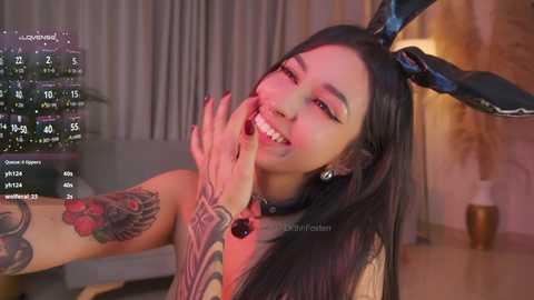 Video of a smiling, tattooed woman with long black hair and bunny ears, wearing a black top, in a dimly lit room with a calendar and window.