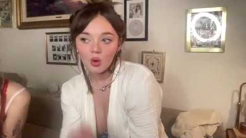 Video of a young woman with fair skin, brown hair, and a surprised expression, wearing a white blouse, pearl necklace, and hoop earrings, in a cozy, framed-wallpapered room with art and a lamp.