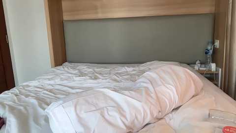 Video of a minimalist bedroom with a neatly made bed, white linens, and a light wood headboard. A bedside table holds a small lamp and a bottle. The room's walls are painted off-white.