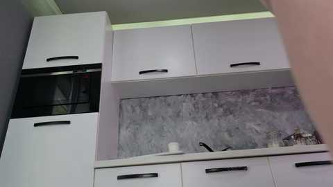 Video of a modern kitchen featuring white cabinets with black handles, a gray marble backsplash, a microwave, a silver knife, and a white container on the counter.