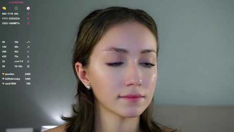 Video of a young woman with fair skin, straight brown hair, and delicate features. She wears a light-colored top. Background includes a muted gray wall with a digital calendar displaying dates and events.